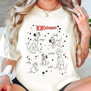 Può includere: T-shirt color crema con la scritta "101 Dalmatians" e illustrazioni dei cani dei cartoni animati Pongo, Lucky, Perdita, Patch e Rolly. Il design include macchie nere e i nomi dei cani.