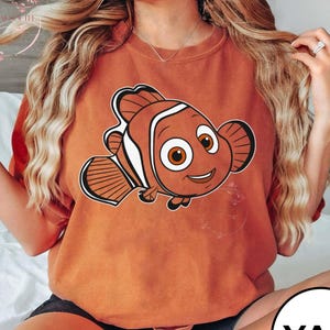 May include: An orange t-shirt featuring a cartoon image of a clownfish. The clownfish has orange, white, and black markings, with large, expressive eyes. The shirt is a solid orange color and appears to be made of a soft material.
