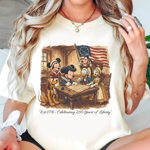 Puede incluir: Camiseta color crema con una ilustración de dibujos animados de personajes de Disney con vestimenta histórica, reunidos alrededor de una mesa con un documento. La bandera estadounidense ondea al fondo. El texto en la parte inferior dice "Est.1776 - Celebrating 250 Years of Liberty."