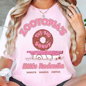 Puede incluir: Camiseta rosa claro con la frase "Zootopia's The Big Donut" en rosa. Un gr&aacute;fico de una rosquilla de chocolate est&aacute; encima de una ilustraci&oacute;n de un cami&oacute;n de comida. El texto "Little Rodentia" y las palabras "Donuts Bagels Coffee" est&aacute;n debajo.