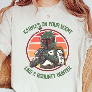 May include: Cream-colored t-shirt featuring a graphic of a bounty hunter with a green and maroon helmet, holding a blaster. The design includes the text "Karma's on Your Scent Like a Bounty Hunter" in green.