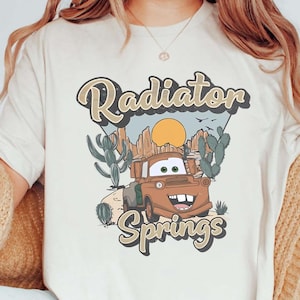 May include: A white t-shirt with a vintage-style graphic of a rusty brown truck with a smiling face. The graphic says "Radiator Springs" with a sunset and cacti in the background.