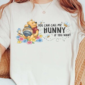 Puede incluir: Camiseta color crema con un gráfico de Winnie the Pooh sosteniendo un tarro de miel, rodeado de flores y abejas. El texto dice "You can call me Hunny if you want".