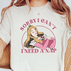 May include: Cream-colored t-shirt featuring a cartoon illustration of a princess with long blonde hair, wearing a pink dress, and resting. The text "SORRY I CAN'T I NEED A NAP" is printed above and below the illustration in pink.