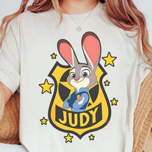 May include: Cream-colored t-shirt featuring a cartoon image of Judy Hopps from Zootopia. Judy is depicted in a police badge with the name "JUDY" below. The design includes yellow stars and a yellow badge.