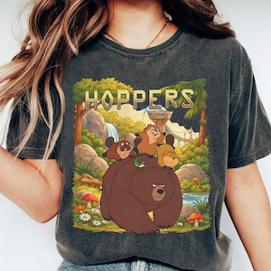 Puede incluir: Camiseta gris oscuro con una ilustración de dibujos animados de un oso y animales más pequeños en un bosque. La palabra "HOPPERS" está encima de la ilustración. El texto "Dark Heather" está en la parte inferior de la camiseta.