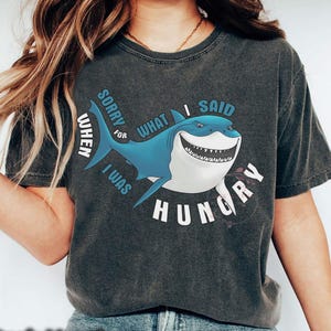 May include: Dark heather gray t-shirt featuring a cartoon shark with the text "Sorry for what I said when I was hungry." The shark is blue and white with a wide grin. The shirt is a casual wear item.