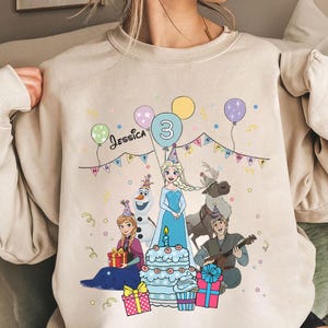 Könnte beinhalten: Beiges Sweatshirt mit einer farbenfrohen Grafik von Frozen-Charakteren, darunter Elsa, Olaf und Kristoff. Das Design zeigt eine Geburtstagstorte, Luftballons und den Namen "Jessica" mit der Zahl "3".