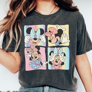 May include: A dark heather grey t-shirt with a graphic of four different images of Minnie Mouse. Each image shows Minnie Mouse with a different expression and pose. The background of the graphic is a rainbow of colors.