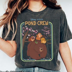 Puede incluir: Camiseta gris oscuro jaspeado con un diseño gráfico de "Pond Crew" de Disney Pixar, que presenta un oso y otros personajes dentro de un marco verde. El texto "Pond Crew" está incluido en el diseño.