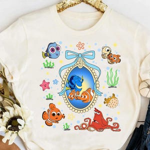 May include: Cream-colored t-shirt featuring a colorful cartoon graphic of characters from the movie "Finding Nemo." The design includes Nemo, Dory, and other sea creatures, surrounded by aquatic elements like seaweed and starfish. A blue ribbon frames the central image.