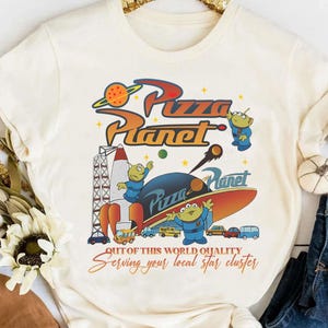 Può includere: T-shirt color crema con grafica colorata del logo Pizza Planet, alieni, un razzo e un'astronave. Il testo recita "Out of this world quality" e "Serving your local star cluster."