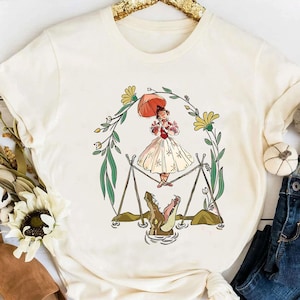 Puede incluir: Camiseta color crema con una ilustración caprichosa. Una mujer con un vestido blanco y detalles rojos se encuentra sobre dos caimanes. El diseño está enmarcado por una corona floral con flores amarillas y hojas verdes. La camiseta es de material suave.