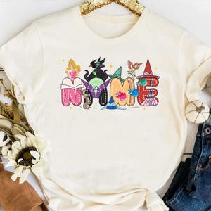 May include: A cream-colored short-sleeve t-shirt featuring a graphic print of the word NAME in large, colorful, stitched-style letters. Each letter incorporates various animated character illustrations, including a princess, a villain, and animals.