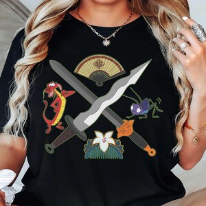 Może przedstawiać: Czarny t-shirt z nadrukiem graficznym przedstawiającym dwa skrzyżowane miecze, czerwonego smoka, fioletowego świerszcza, ozdobny składany wachlarz oraz biały grzebień do włosów w kształcie kwiatu, umieszczonym centralnie na klatce piersiowej.
