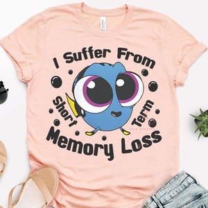 May include: A light pink t-shirt featuring a cartoon image of Dory from Finding Nemo. The blue fish has large eyes and is surrounded by the text "I Suffer From Short Term Memory Loss."