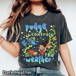 Puede incluir: Camiseta gris oscuro con un gr&aacute;fico colorido que dice "Pudge controls the weather". El dise&ntilde;o incorpora personajes de dibujos animados y motivos acu&aacute;ticos. Esta camiseta es una prenda casual.