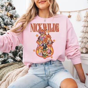 May include: A light pink sweatshirt featuring a graphic of Nick Wilde and other characters from Zootopia. The text "NICK WILDE" is arched above the characters in a retro font. The sweatshirt is long-sleeved and has a crew neck.