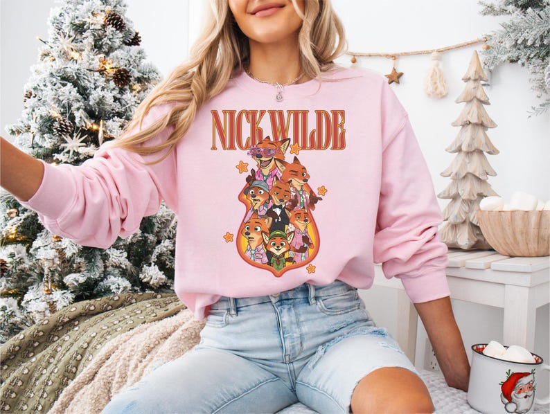 May include: A light pink sweatshirt featuring a graphic of Nick Wilde and other characters from Zootopia. The text "NICK WILDE" is arched above the characters in a retro font. The sweatshirt is long-sleeved and has a crew neck.