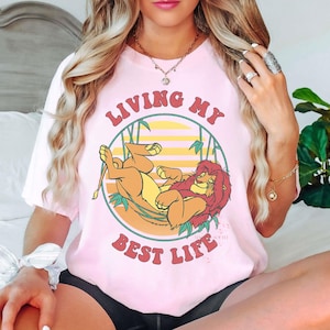 May include: A light pink t-shirt featuring a cartoon illustration of Simba and Mufasa from The Lion King, with the text "LIVING MY BEST LIFE" in a circular design. The design includes a yellow and orange sunset background.