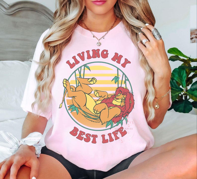May include: A light pink t-shirt featuring a cartoon illustration of Simba and Mufasa from The Lion King, with the text "LIVING MY BEST LIFE" in a circular design. The design includes a yellow and orange sunset background.