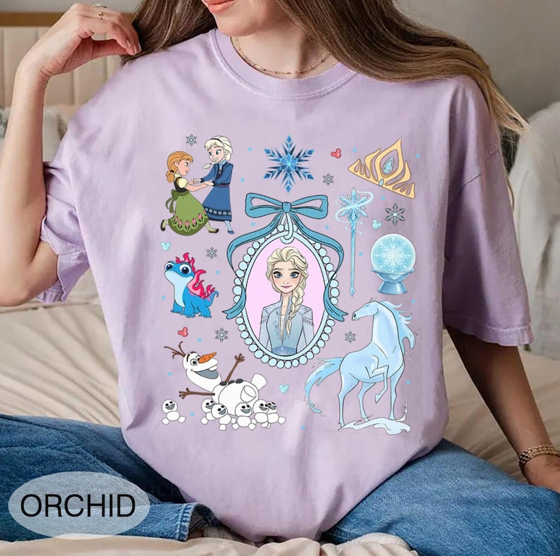May include: A lilac t-shirt with a cartoon design featuring characters and symbols from a popular animated film. The design includes images of two princesses, a snowman, a dragon, and a horse. The word "ORCHID" is printed at the bottom.