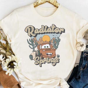 May include: A white t-shirt with a vintage-style graphic of a rusty brown truck with a smiling face. The graphic says "Radiator Springs" in a retro font.