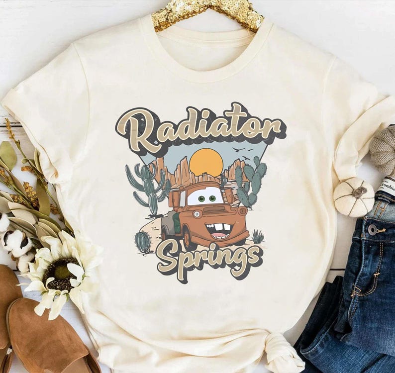 May include: A white t-shirt with a vintage-style graphic of a rusty brown truck with a smiling face. The graphic says "Radiator Springs" in a retro font.