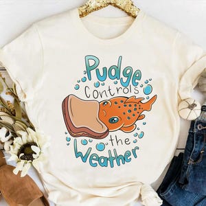 May include: A white t-shirt with a cartoon illustration of a fish eating a peanut butter and jelly sandwich. The text "Pudge controls the weather" is printed on the shirt.