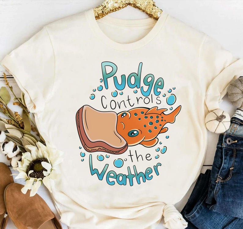 May include: A white t-shirt with a cartoon illustration of a fish eating a peanut butter and jelly sandwich. The text "Pudge controls the weather" is printed on the shirt.