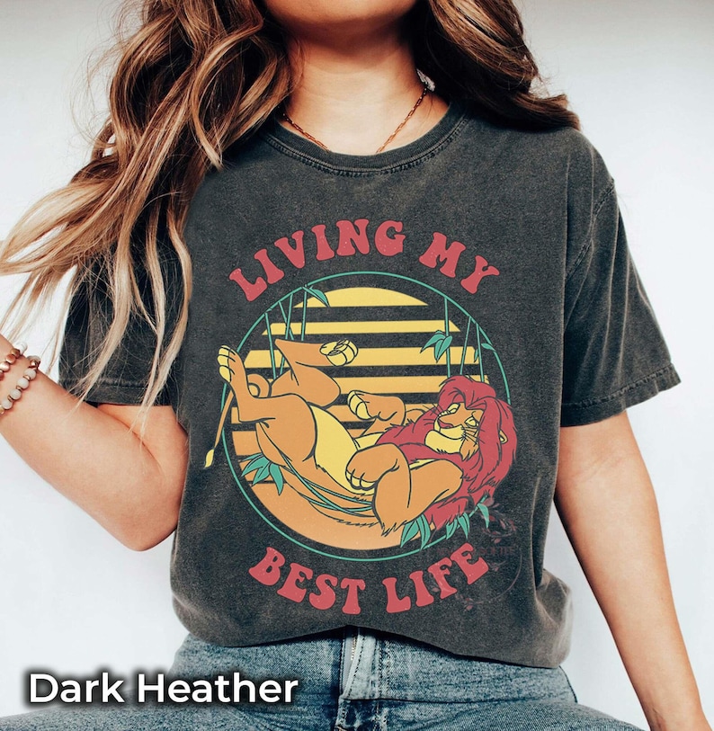 May include: Dark heather gray t-shirt featuring a cartoon lion lounging in a circle with the text "LIVING MY BEST LIFE" in red and orange. The shirt is a relaxed fit.