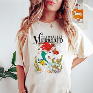 Op de afbeelding: Een wit T-shirt met een afbeelding van Ariel, de Kleine Zeemeermin, die zwemt met Flounder. De tekst "The Little Mermaid" is boven de afbeelding gedrukt.