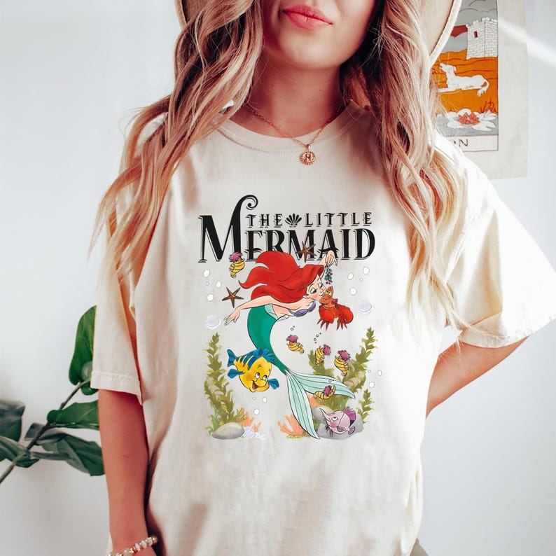 Op de afbeelding: Een wit T-shirt met een afbeelding van Ariel, de Kleine Zeemeermin, die zwemt met Flounder. De tekst "The Little Mermaid" is boven de afbeelding gedrukt.