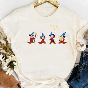 May include: Cream-colored t-shirt featuring a cartoon design of Mickey Mouse in a wizard outfit. The design shows Mickey in four different poses, each holding a different magical item. The shirt has a round neckline and long sleeves.