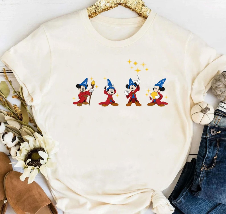 May include: Cream-colored t-shirt featuring a cartoon design of Mickey Mouse in a wizard outfit. The design shows Mickey in four different poses, each holding a different magical item. The shirt has a round neckline and long sleeves.
