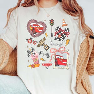 May include: Cream-colored t-shirt featuring a colorful graphic design of the Cars movie. The design includes images of Lightning McQueen, racing flags, and other related elements. The word "Cars" is also printed on the shirt.