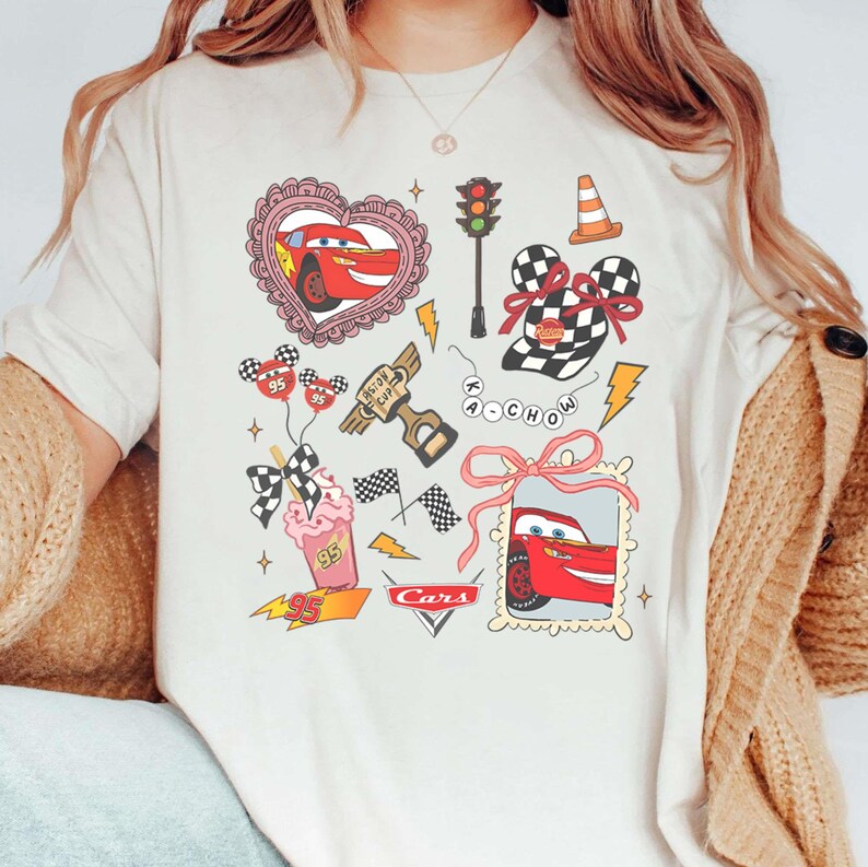May include: Cream-colored t-shirt featuring a colorful graphic design of the Cars movie. The design includes images of Lightning McQueen, racing flags, and other related elements. The word "Cars" is also printed on the shirt.
