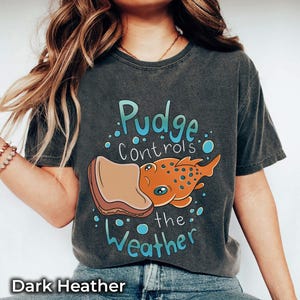 May include: A dark heather grey t-shirt with a cartoon illustration of a fish with a slice of bread on its head. The text "Pudge controls the weather" is printed on the shirt.