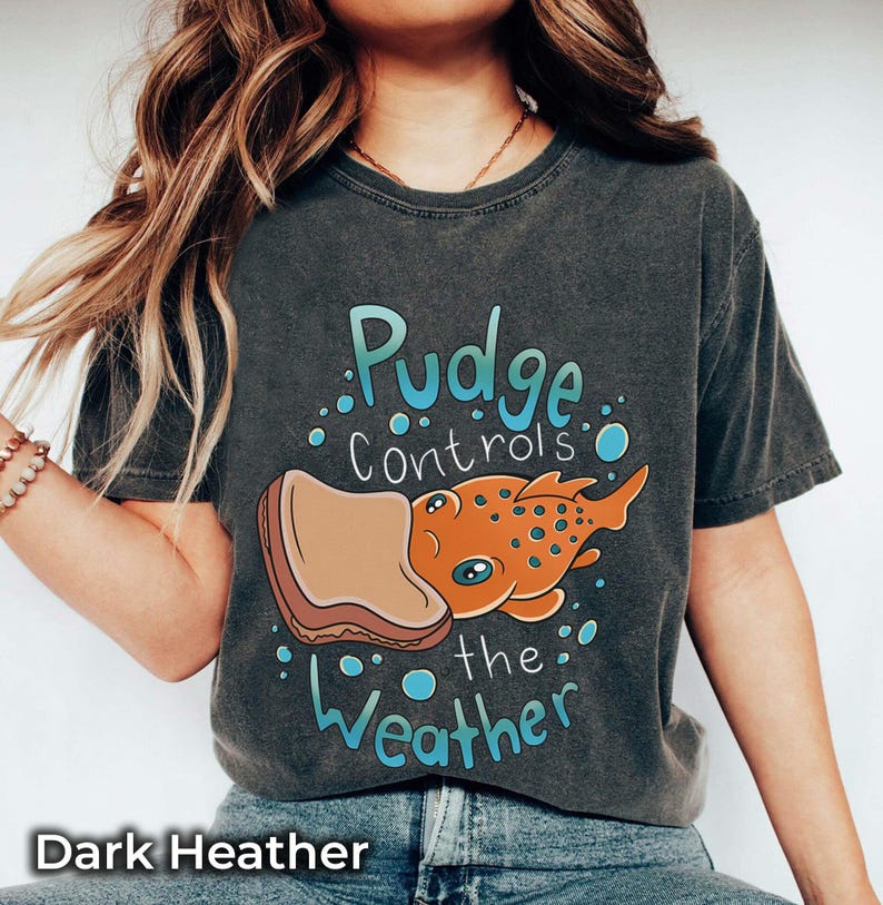 May include: A dark heather grey t-shirt with a cartoon illustration of a fish with a slice of bread on its head. The text "Pudge controls the weather" is printed on the shirt.
