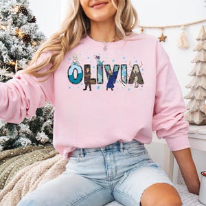 Pu&ograve; includere: Una felpa rosa chiaro con il nome "OLIVIA" stampato sul davanti in un carattere colorato e a cartoni animati. Le lettere sono decorate con immagini di personaggi dei cartoni animati. La felpa &egrave; a maniche lunghe e ha uno scollo tondo.