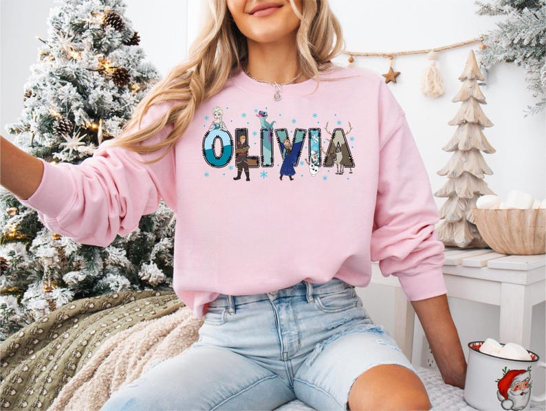 Pu&ograve; includere: Una felpa rosa chiaro con il nome "OLIVIA" stampato sul davanti in un carattere colorato e a cartoni animati. Le lettere sono decorate con immagini di personaggi dei cartoni animati. La felpa &egrave; a maniche lunghe e ha uno scollo tondo.