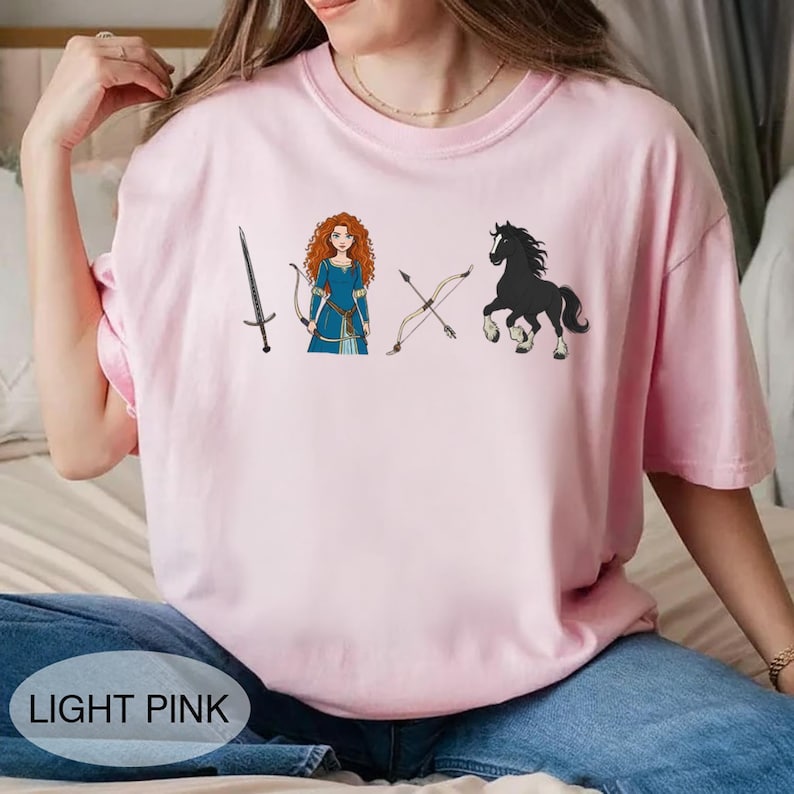 May include: Light pink t-shirt featuring a graphic of a sword, a woman in a blue dress, crossed bows, and a black horse. The text "LIGHT PINK" is at the bottom. The shirt is made of a soft material.