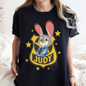 May include: Black t-shirt featuring a cartoon image of Judy Hopps from Zootopia. The design includes Judy in a police uniform, set against a yellow badge with the name "JUDY" and yellow stars.