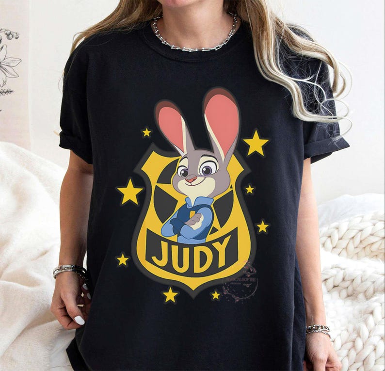 May include: Black t-shirt featuring a cartoon image of Judy Hopps from Zootopia. The design includes Judy in a police uniform, set against a yellow badge with the name "JUDY" and yellow stars.