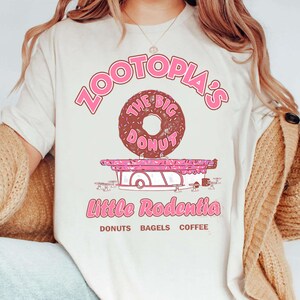 Puede incluir: Camiseta color crema con un gr&aacute;fico rosa de una tienda de donuts de Zootopia. El dise&ntilde;o incluye el texto "Zootopia's The Big Donut" encima de una rosquilla marr&oacute;n y "Little Rodentia" debajo, con "Donuts Bagels Coffee" en la parte inferior.