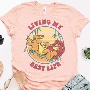 May include: A light pink t-shirt featuring a cartoon lion design within a circular frame. The lion is depicted in shades of orange and yellow, with a red mane. The text "LIVING MY BEST LIFE" is arched above and below the lion.