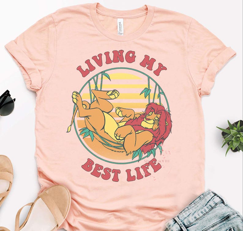 May include: A light pink t-shirt featuring a cartoon lion design within a circular frame. The lion is depicted in shades of orange and yellow, with a red mane. The text "LIVING MY BEST LIFE" is arched above and below the lion.