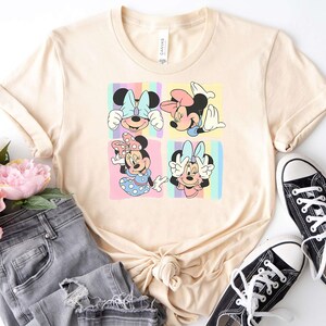 May include: A light beige t-shirt with a graphic print of four different images of Minnie Mouse. Each image shows Minnie Mouse with a different expression and pose. The background of each image is a pastel rainbow.