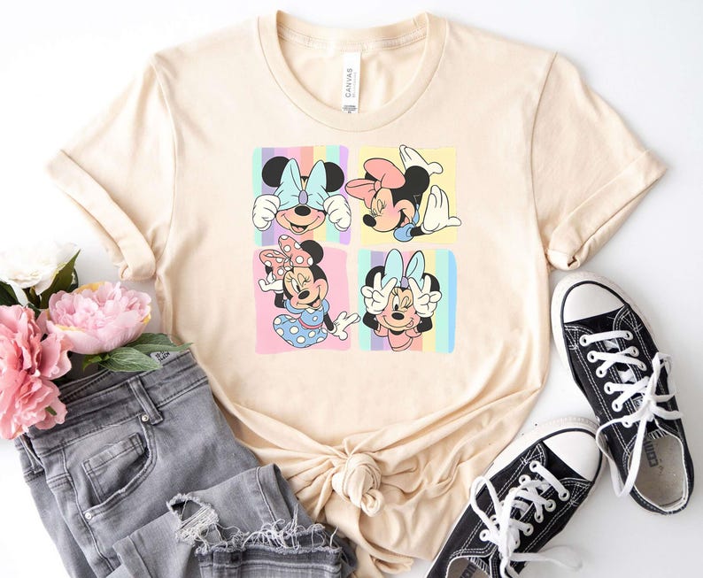 May include: A light beige t-shirt with a graphic print of four different images of Minnie Mouse. Each image shows Minnie Mouse with a different expression and pose. The background of each image is a pastel rainbow.