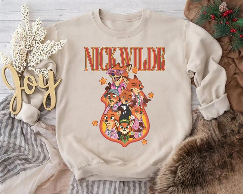 May include: Beige crewneck sweatshirt featuring a graphic of Nick Wilde and other characters from Zootopia. The text "NICK WILDE" is in orange and red lettering above the characters. The design includes stars and a cartoon style.